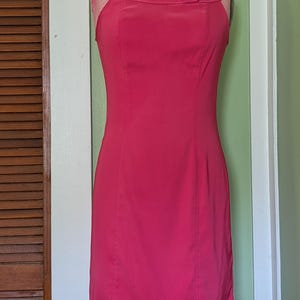 May include: A sleeveless, knee-length, fuchsia-colored dress displayed on a vintage dress form. The dress features a unique neckline with a small button detail and a fitted silhouette. The dress is made of a smooth fabric and has vertical seams.