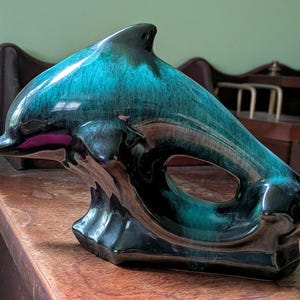 May include: A ceramic dolphin figurine with a glossy, teal and black finish. The dolphin is posed in a leaping position, with a curved body and a fin. The figurine is a decorative item.