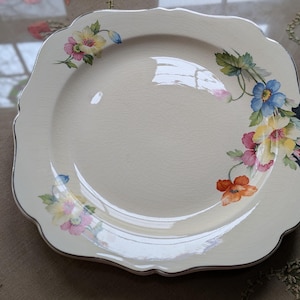 May include: Cream-colored square plate with a scalloped edge and a silver trim. The plate is decorated with colorful floral designs in shades of pink, yellow, blue, and orange. A vintage piece for serving.
