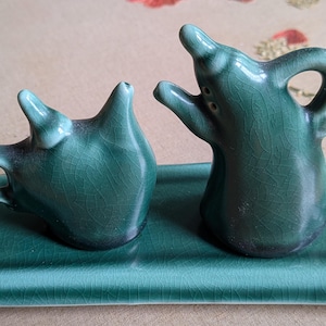 May include: A set of two green ceramic salt and pepper shakers with a crackle glaze finish, each featuring a handle and a spout-like top, resting on a matching rectangular green ceramic tray with a consistent crackle glaze texture throughout.