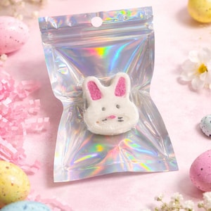 May include: A white bunny-shaped bath bomb with pink ears and facial details is packaged in a holographic, iridescent zip-lock bag. The Easter-themed image includes pastel-colored eggs, flowers, and pink paper shreds on a light pink surface.