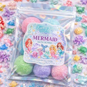 May include: A clear plastic bag filled with colorful bath bombs, labeled "Mystery Toy Mermaid Bath Bombs" by Faith & Fizz Soap Co. The bag is surrounded by mermaid-themed figurines and glittery embellishments in various colors, including pink, blue, and purple.