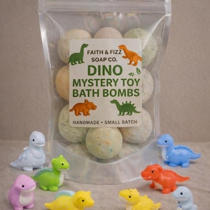 May include: A clear bag of Dino Mystery Toy Bath Bombs from Faith & Fizz Soap Co. is surrounded by colorful toy dinosaurs. The bath bombs are round and speckled. The bag has a label with dinosaur illustrations and the words "Handmade Small Batch."