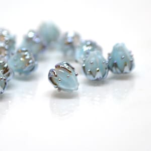 May include: A group of light blue glass beads with silver accents. The beads are shaped like a flower bud and have an iridescent finish.