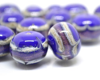 Puede incluir: Primer plano de varias cuentas de vidrio azul con un acabado brillante. Cada cuenta presenta un patrón único de remolinos blancos y morados, con un toque de purpurina dorada. Las cuentas son redondas con un agujero para ensartar.