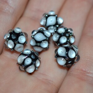 May include: A collection of six black and white glass beads with a textured surface. The beads are round and have a speckled pattern.