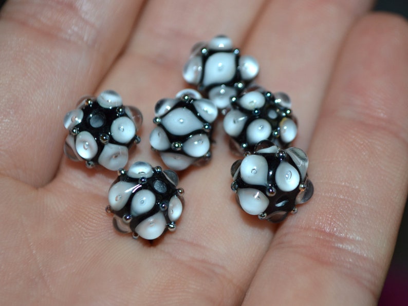 May include: A collection of six black and white glass beads with a textured surface. The beads are round and have a speckled pattern.