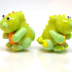 May include: Two green glass dinosaur beads with orange accents. The beads are hand-blown and have a unique, whimsical design.