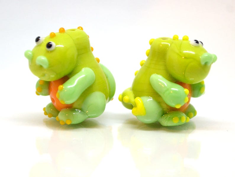 May include: Two green glass dinosaur beads with orange accents. The beads are hand-blown and have a unique, whimsical design.