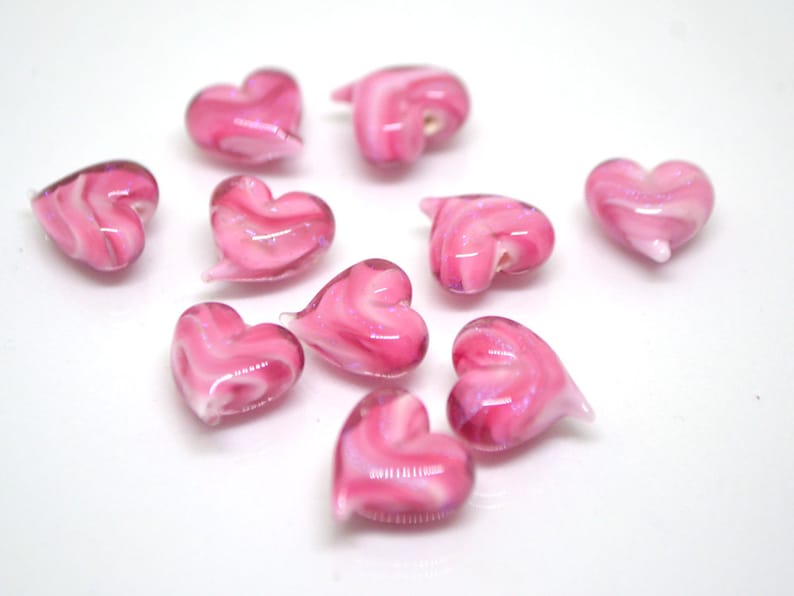 Peut inclure: Dix perles en verre en forme de c&oelig;ur de couleur rose avec des tourbillons blancs. Les perles sont &eacute;parpill&eacute;es sur une surface blanche.