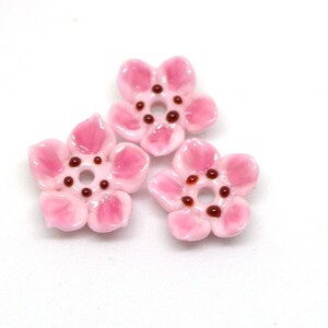 May include: Three pink ceramic flower beads with brown centers. The beads are perfect for jewelry making or crafting.