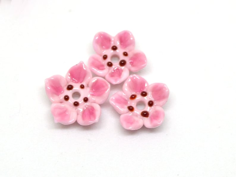 May include: Three pink ceramic flower beads with brown centers. The beads are perfect for jewelry making or crafting.
