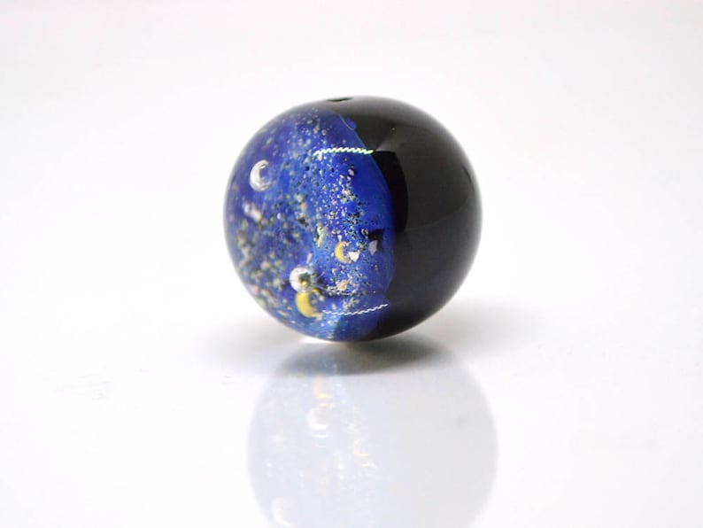 May include: A dark blue glass bead with a galaxy design. The bead is filled with gold and white glitter and has a black background.