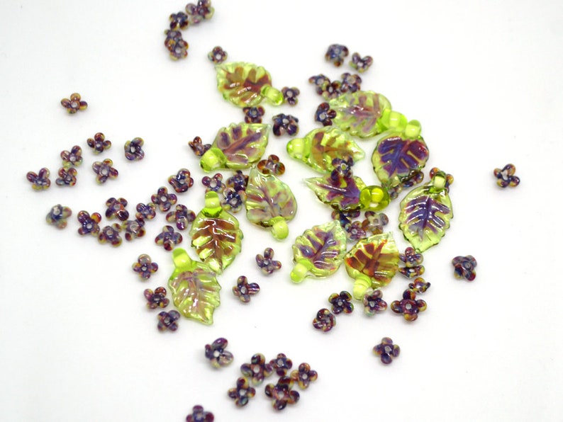 May include: Assortment of green and purple glass beads in the shape of leaves and flowers. The beads are iridescent and have a glossy finish.