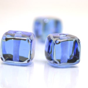 Puede incluir: Tres cuentas de vidrio cúbicas azules con un centro transparente. Las cuentas son facetadas y tienen un ligero brillo iridiscente.