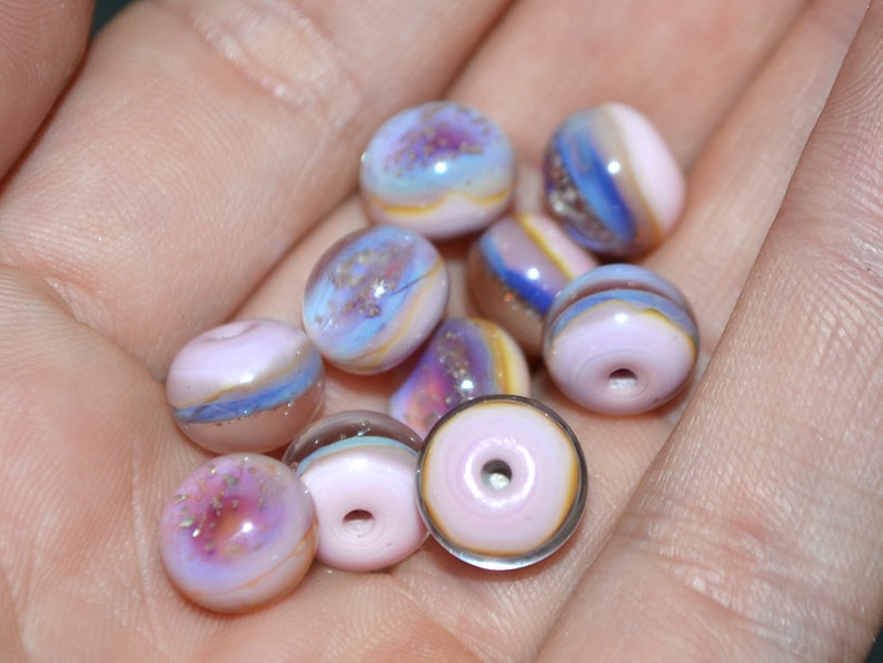 May include: A collection of 10 small, round, glass beads in shades of pink, purple, and blue with iridescent glitter. The beads have a layered, marbled effect.