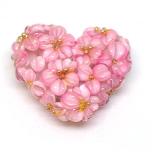 May include: A pink glass heart-shaped charm with a floral design. The charm is made of small pink glass beads that are arranged to look like flowers. The flowers have gold centers.