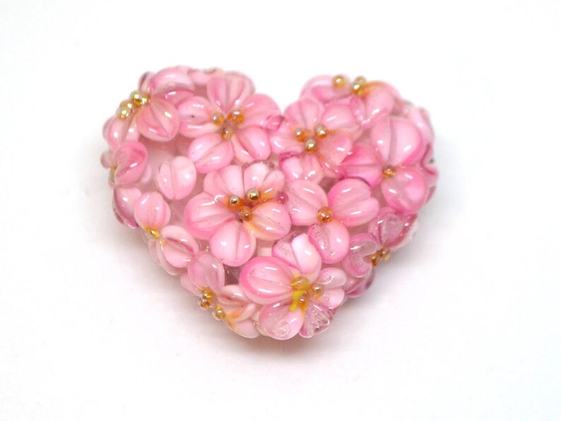 May include: A pink glass heart-shaped charm with a floral design. The charm is made of small pink glass beads that are arranged to look like flowers. The flowers have gold centers.