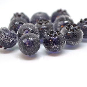 May include: Close-up of a group of dark blue glass blueberries with a frosted finish. The blueberries are arranged in a cluster on a white surface.