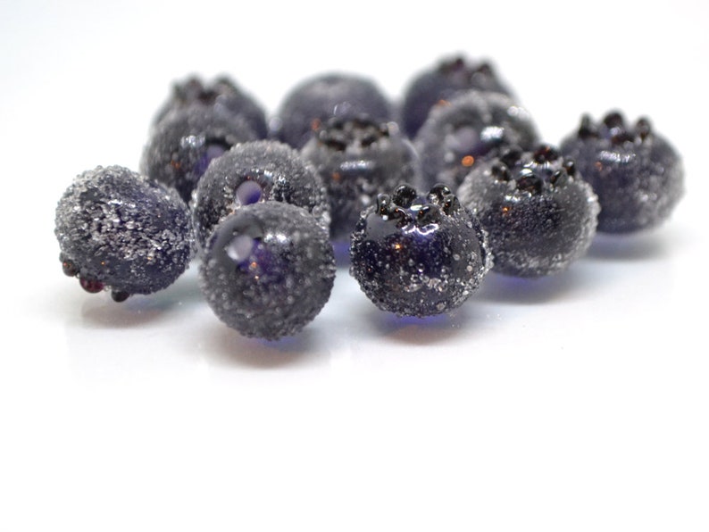 May include: Close-up of a group of dark blue glass blueberries with a frosted finish. The blueberries are arranged in a cluster on a white surface.