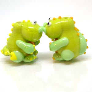 May include: Two green glass dinosaur figurines with yellow and orange accents. The dinosaurs are facing each other and appear to be kissing.
