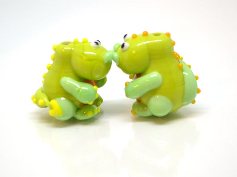 May include: Two green glass dinosaur figurines with yellow and orange accents. The dinosaurs are facing each other and appear to be kissing.