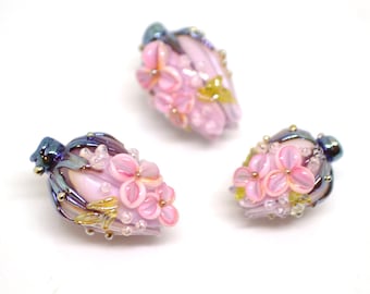 May include: Three glass beads with pink and white flowers and iridescent blue and gold accents.