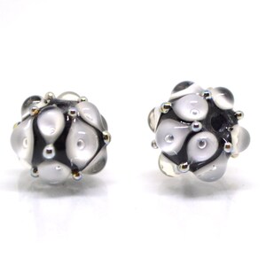 May include: Two black and white glass beads with a textured surface and silver accents. The beads are round and have a cluster of white circles on the surface.
