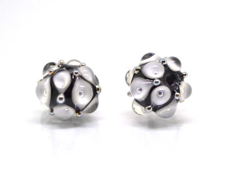 May include: Two black and white glass beads with a textured surface and silver accents. The beads are round and have a cluster of white circles on the surface.