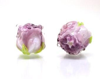 Könnte beinhalten: Zwei Glasperlen in Form von Rosen. Die Perlen sind weiß mit violetten Wirbeln und grünen Akzenten.
