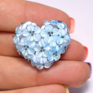 May include: A heart-shaped charm made of blue and white glass beads, resembling a cluster of delicate flowers. Each flower has a white center and a blue outer layer, with small silver beads at the center of each flower.