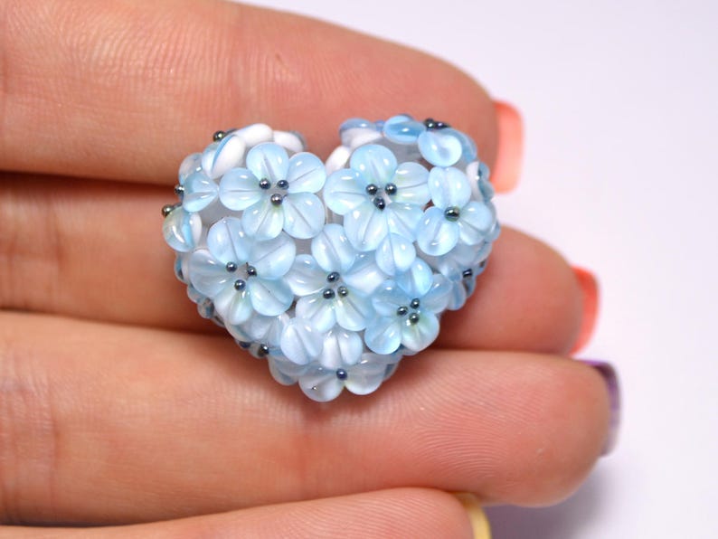 May include: A heart-shaped charm made of blue and white glass beads, resembling a cluster of delicate flowers. Each flower has a white center and a blue outer layer, with small silver beads at the center of each flower.