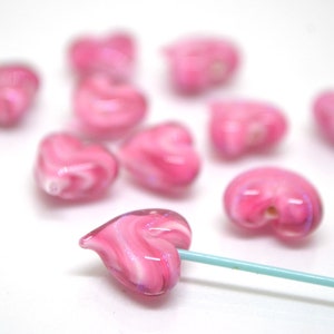 Peut inclure: Un ensemble de perles en verre en forme de c&oelig;ur rose avec des tourbillons de blanc. Les perles sont &eacute;parpill&eacute;es sur une surface blanche. Une perle est enfil&eacute;e sur une ficelle turquoise.