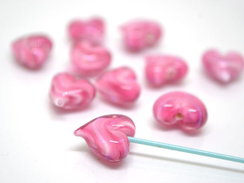 Peut inclure: Un ensemble de perles en verre en forme de c&oelig;ur rose avec des tourbillons de blanc. Les perles sont &eacute;parpill&eacute;es sur une surface blanche. Une perle est enfil&eacute;e sur une ficelle turquoise.