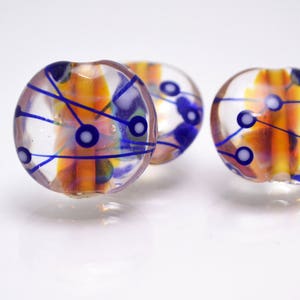 Pu&ograve; includere: Tre perline di vetro con una base trasparente e disegni astratti blu e arancioni. Le perline hanno punti bianchi e linee blu.
