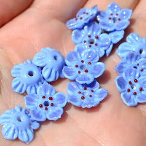 May include: A collection of light blue glass flower beads with red centers. The beads are arranged in a hand, with the centers of the flowers facing up.