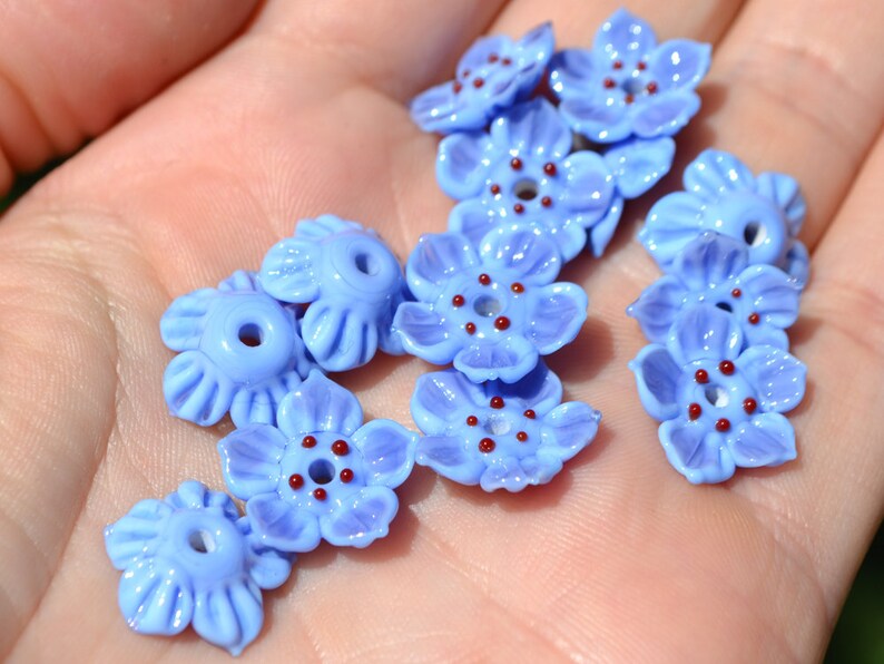 May include: A collection of light blue glass flower beads with red centers. The beads are arranged in a hand, with the centers of the flowers facing up.