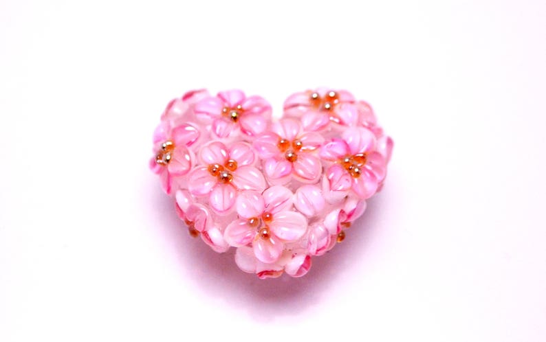 May include: A pink heart-shaped ornament made of glass beads, resembling a cluster of delicate pink flowers with gold accents.