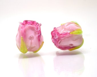Könnte beinhalten: Zwei rosa Glasrosenperlen mit grünen Akzenten. Die Perlen sind aus Glas und haben eine realistische Rosenform.
