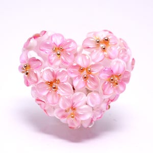 May include: A pink heart-shaped ornament made of glass beads, resembling a cluster of cherry blossoms. Each flower has a delicate pink hue and a gold center.