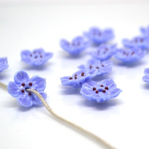 May include: A collection of light blue glass flower beads with maroon centers. The beads are strung on a white cord.