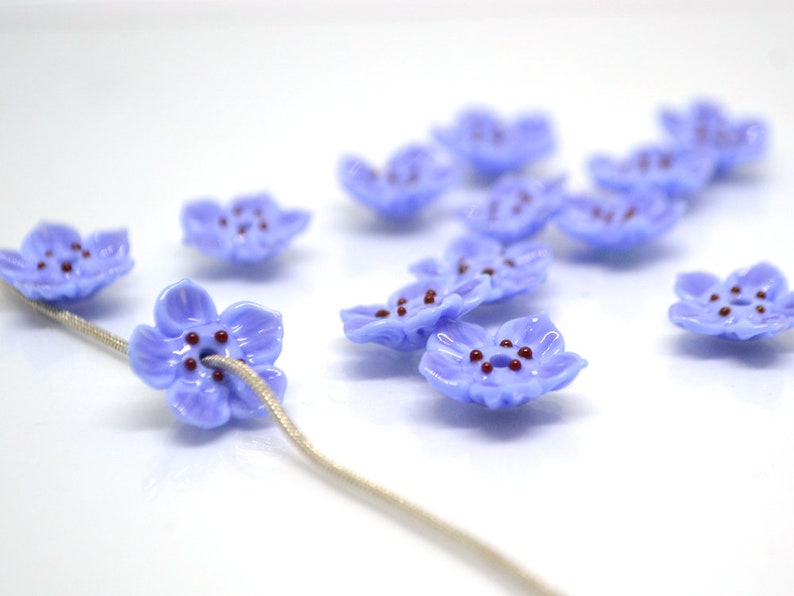 May include: A collection of light blue glass flower beads with maroon centers. The beads are strung on a white cord.