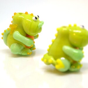 May include: Two green glass dinosaur figurines with yellow accents. The dinosaurs are holding small orange balls in their hands.