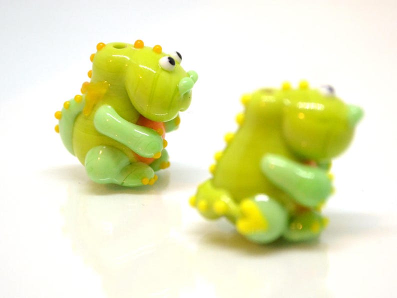 May include: Two green glass dinosaur figurines with yellow accents. The dinosaurs are holding small orange balls in their hands.