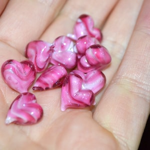Peut inclure: Un ensemble de perles de verre en forme de c&oelig;ur rose et blanc avec des tourbillons de couleur. Les perles sont dans la main d'une personne.