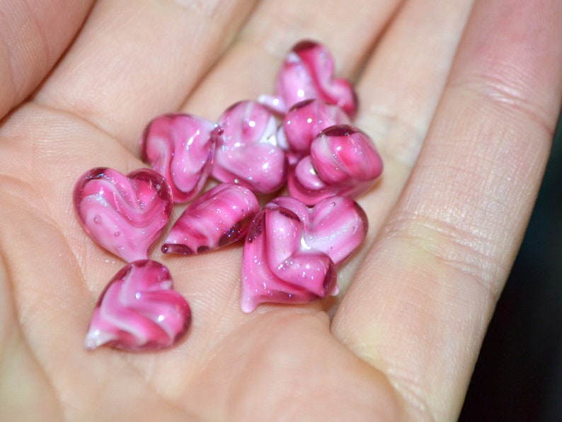 Peut inclure: Un ensemble de perles de verre en forme de c&oelig;ur rose et blanc avec des tourbillons de couleur. Les perles sont dans la main d'une personne.