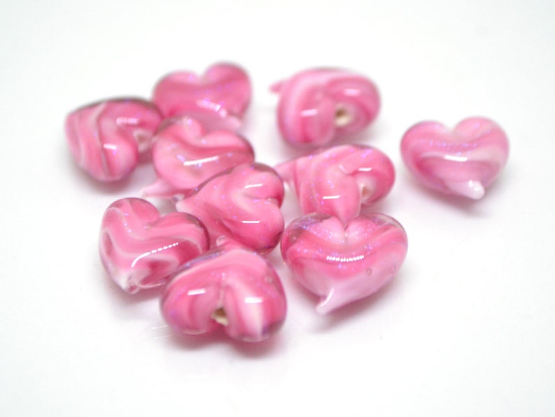 Peut inclure: Un ensemble de perles en verre en forme de c&oelig;ur de couleur rose avec des tourbillons blancs. Les perles sont de tailles et de formes diff&eacute;rentes et ont une finition brillante.