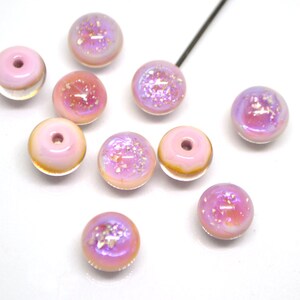 May include: Ten pink iridescent glass beads with a hole in the center. The beads have a light pink base with a clear iridescent coating and white glitter.