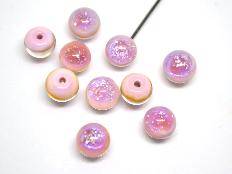 May include: Ten pink iridescent glass beads with a hole in the center. The beads have a light pink base with a clear iridescent coating and white glitter.