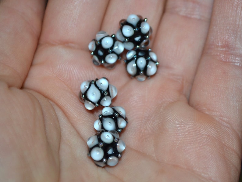 May include: Five white and black glass beads with a textured surface. The beads are round and have a cluster of small white dots on each bead.